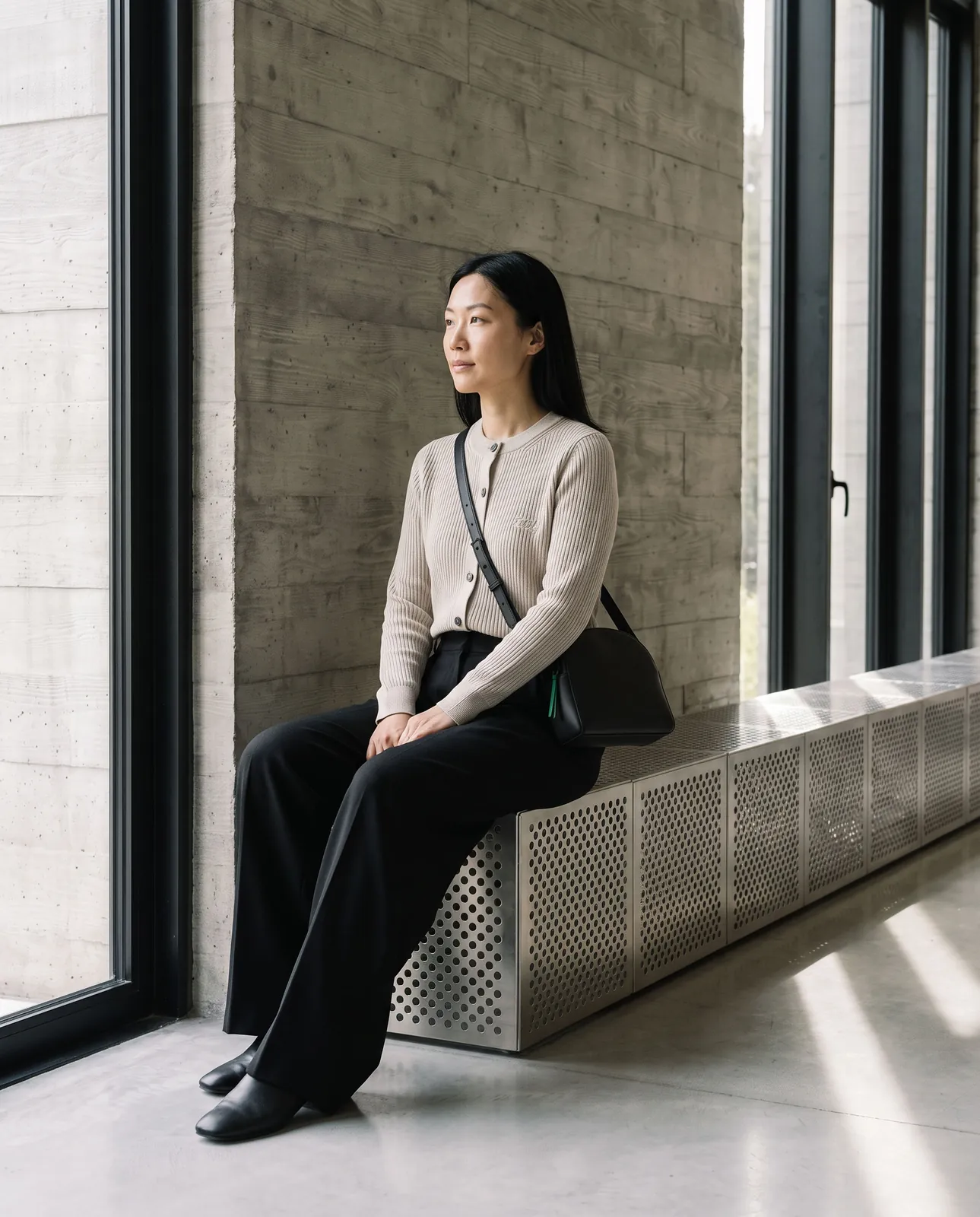 A woman in the oat cardigan sits on the perforated steel bench, crossbody at her side